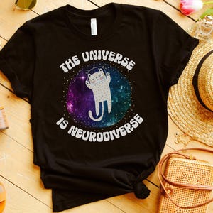 Puede incluir: Camiseta negra con el texto blanco "THE UNIVERSE IS NEURODIVERSE", que rodea un gato de dibujos animados en un diseño de galaxia. La camiseta se muestra sobre una superficie de madera con sandalias, un sombrero de paja y otros accesorios.
