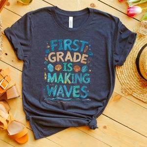 Pode incluir: Uma camiseta azul escura com o texto "First Grade is Making Waves" em letras coloridas. O texto &eacute; cercado por imagens de conchas, um farol e ondas.