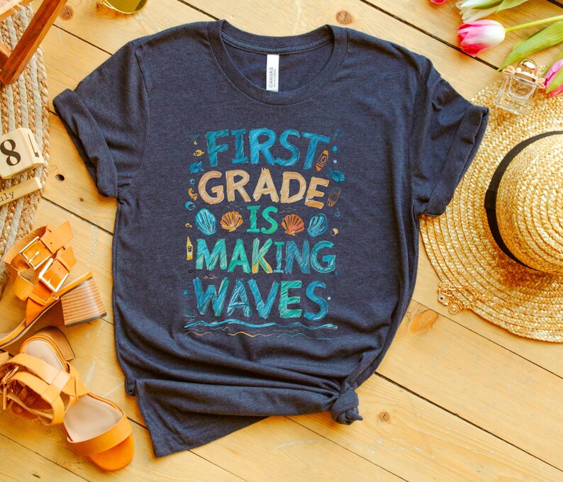 Pode incluir: Uma camiseta azul escura com o texto "First Grade is Making Waves" em letras coloridas. O texto &eacute; cercado por imagens de conchas, um farol e ondas.