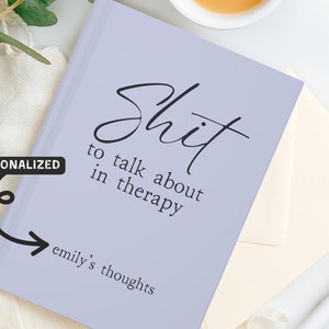 Op de afbeelding: Een lila notitieboek met de tekst "Shit to talk about in therapy" in zwart schrift. Het notitieboek bevat ook de tekst "emily's thoughts". Een gepersonaliseerd cadeau-idee.