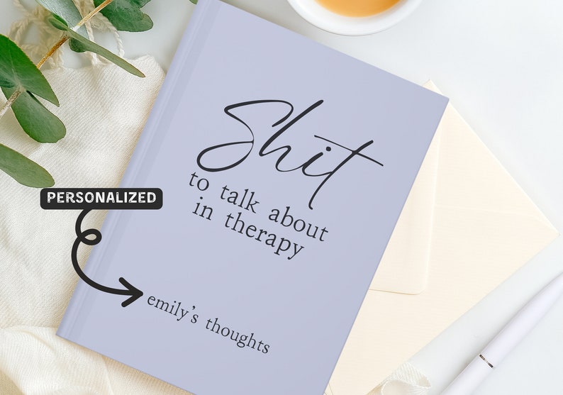 Op de afbeelding: Een lila notitieboek met de tekst "Shit to talk about in therapy" in zwart schrift. Het notitieboek bevat ook de tekst "emily's thoughts". Een gepersonaliseerd cadeau-idee.