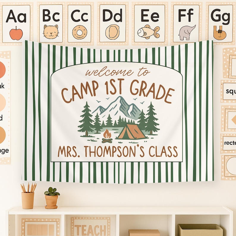 May include: A white and green striped banner with the text "Welcome to Camp 1st Grade" and "Mrs. Thompson's Class". The banner features a cartoon illustration of a campsite with a tent, mountains, trees, and a campfire.