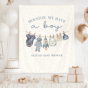 May include: A baby shower banner with the text "Houston, we have a boy" and "Olivia's Baby Shower". The banner features illustrations of baby clothes and space-themed elements in blue and white.
