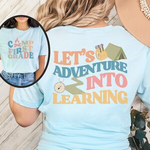 May include: A light blue t-shirt with a camping theme. The front of the shirt says "Camp First Grade" with a campfire graphic. The back of the shirt says "Let's Adventure Into Learning" with a compass, a hiking trail, a backpack, and a tent graphic.