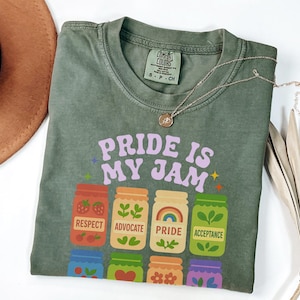 Puede incluir: Una camiseta verde oliva con el texto "PRIDE IS MY JAM" en morado. Debajo del texto hay ilustraciones de tarros de mermelada con las palabras "RESPECT", "ADVOCATE", "PRIDE" y "ACCEPTANCE". Un collar dorado está sobre la camiseta.