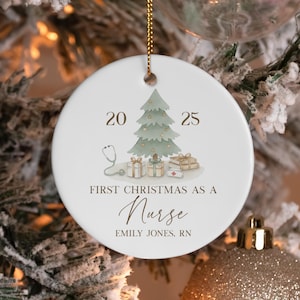 Könnte beinhalten: Ein runder, weißer Keramik-Weihnachtsbaumschmuck mit einer Aquarellillustration eines Weihnachtsbaums, Geschenken, einem Stethoskop und einer Krankenschwesterhaube. Der Text lautet 20 25, FIRST CHRISTMAS AS A Nurse und EMILY JONES, RN. Er hängt an einer goldenen Schnur.