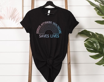 May include: Black t-shirt with a rainbow graphic and the text "GENDER-AFFIRMING HEALTHCARE SAVES LIVES". The shirt is hanging on a wooden ladder against a white wall. A framed picture and green plant are in the background.