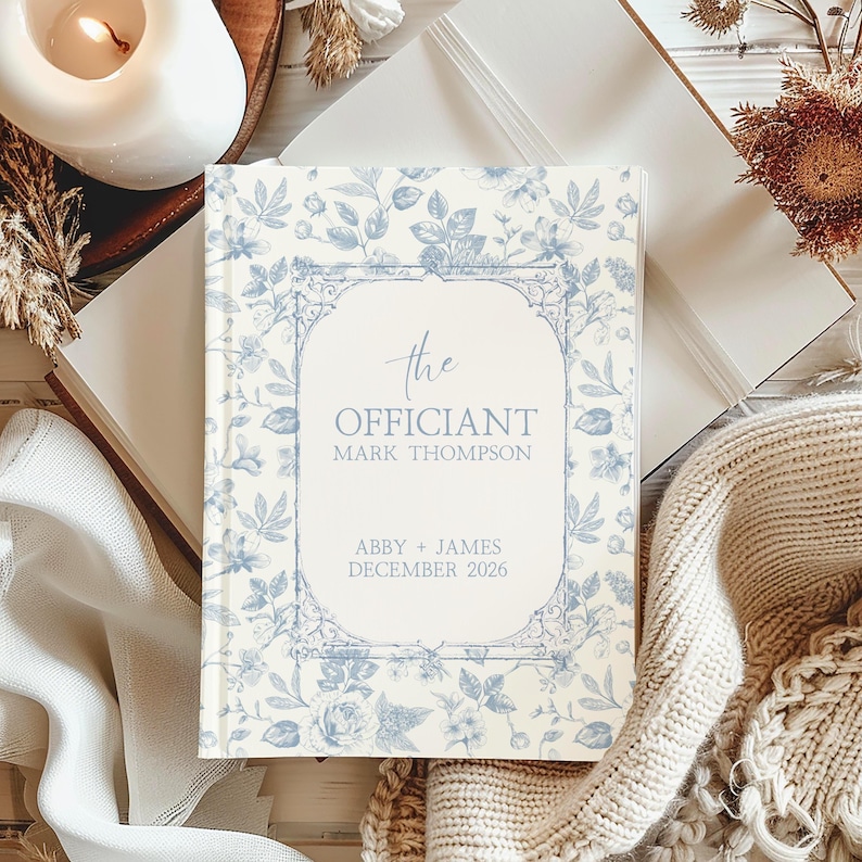 Puede incluir: Un libro de tapa dura con un estampado floral en azul claro y blanco. La portada presenta el texto "the Officiant" y los nombres Mark Thompson, Abby & James, y Diciembre 2026. El libro est&aacute; rodeado de elementos decorativos.