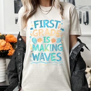 Pode incluir: Uma camiseta bege claro com o texto "First Grade is Making Waves" em letras coloridas desenhadas &agrave; m&atilde;o. O texto &eacute; rodeado por ilustra&ccedil;&otilde;es de conchas e ondas.