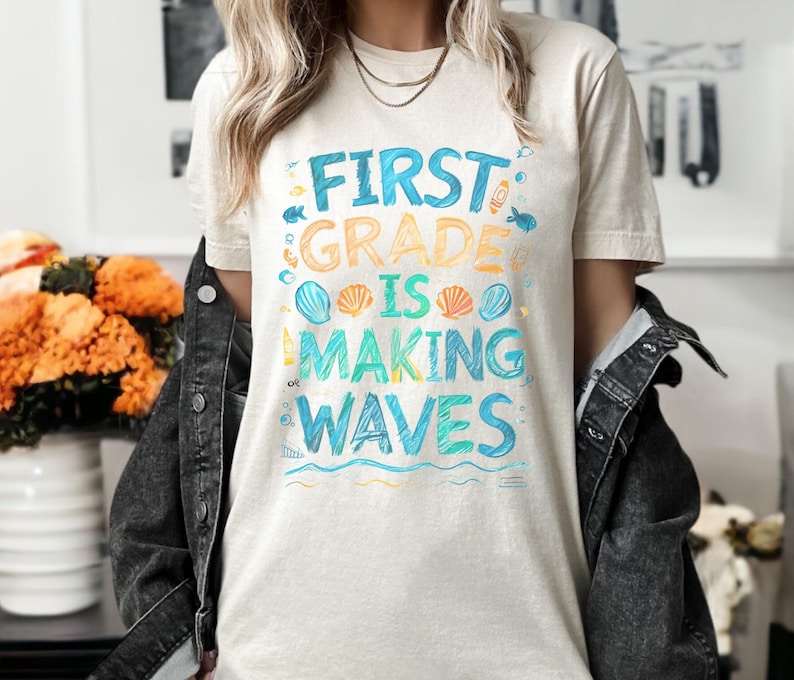 Pode incluir: Uma camiseta bege claro com o texto "First Grade is Making Waves" em letras coloridas desenhadas &agrave; m&atilde;o. O texto &eacute; rodeado por ilustra&ccedil;&otilde;es de conchas e ondas.