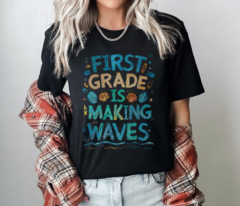 Pode incluir: Camiseta preta com o texto "First Grade is Making Waves" em letras coloridas. O texto &eacute; rodeado por ilustra&ccedil;&otilde;es de conchas, l&aacute;pis e um farol.