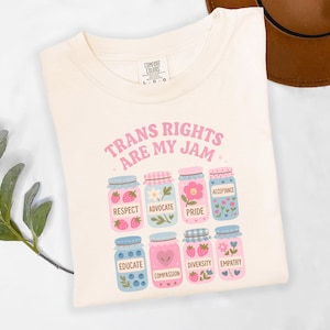 Może przedstawiać: Kremowa koszulka z nadrukiem "TRANS RIGHTS ARE MY JAM" w kolorze różowym. Tekst jest otoczony ilustracjami słoików z dżemem z różnymi napisami na każdym słoiku, w tym "RESPECT", "ADVOCATE", "PRIDE", "ACCEPTANCE", "EDUCATE", "COMPASSION", "DIVERSITY" i "EMPATHY".