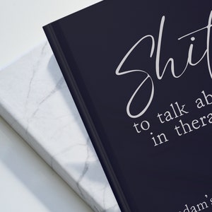 Op de afbeelding: Twee hardcover boeken. E&eacute;n is zwart met witte tekst die luidt: "Shit to talk about in therapy". Het andere boek is wit met een marmerpatroon. De boeken staan schuin naar elkaar toe.