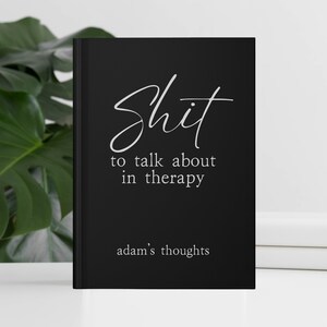 Op de afbeelding: Een zwart notitieboek met witte tekst "Shit to talk about in therapy". Het notitieboek bevat ook de tekst "adam's thoughts" onderaan. Het notitieboek ligt op een wit oppervlak met een groene plant op de achtergrond.
