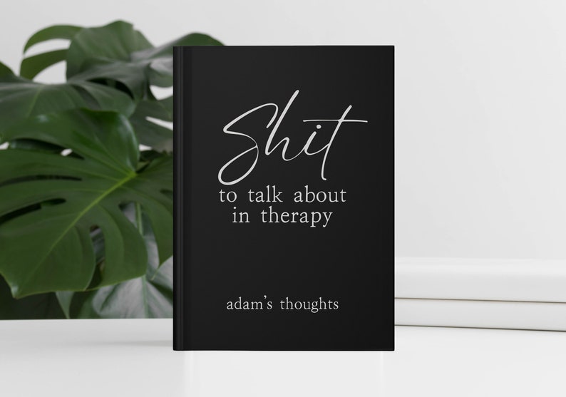 Op de afbeelding: Een zwart notitieboek met witte tekst "Shit to talk about in therapy". Het notitieboek bevat ook de tekst "adam's thoughts" onderaan. Het notitieboek ligt op een wit oppervlak met een groene plant op de achtergrond.