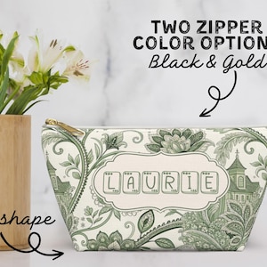K&ouml;nnte beinhalten: Eine T-f&ouml;rmige Kosmetiktasche mit goldenem Rei&szlig;verschluss, mit einem gr&uuml;n-wei&szlig;en Blumen- und Architekturdesign. Der Name "LAURIE" ist auf der Vorderseite abgebildet. Der Text oben lautet "TWO ZIPPER COLOR OPTIONS Black & Gold."