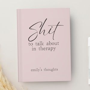 Op de afbeelding: Een lichtroze notitieboek met de tekst "Shit to talk about in therapy" in zwart script. De tekst "emily's thoughts" staat eronder. Rechts van het notitieboek ligt een witte pen.