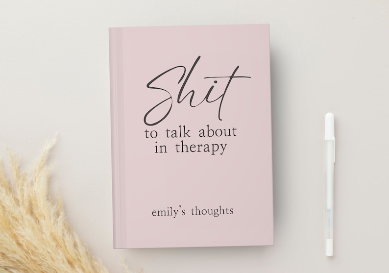 Op de afbeelding: Een lichtroze notitieboek met de tekst "Shit to talk about in therapy" in zwart script. De tekst "emily's thoughts" staat eronder. Rechts van het notitieboek ligt een witte pen.