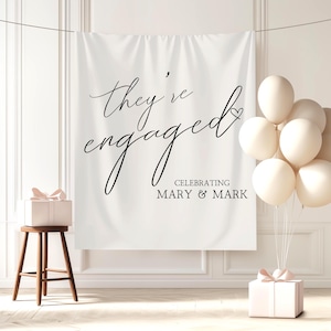May include: A white fabric backdrop with the text "they're engaged" in elegant black script, celebrating Mary & Mark. A small wooden stool holds a gift box with a pink bow. A cluster of cream-colored balloons and another gift box complete the scene.