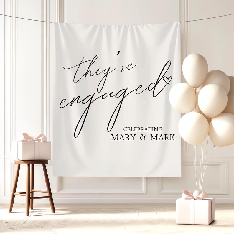 May include: A white fabric backdrop with the text "they're engaged" in elegant black script, celebrating Mary & Mark. A small wooden stool holds a gift box with a pink bow. A cluster of cream-colored balloons and another gift box complete the scene.