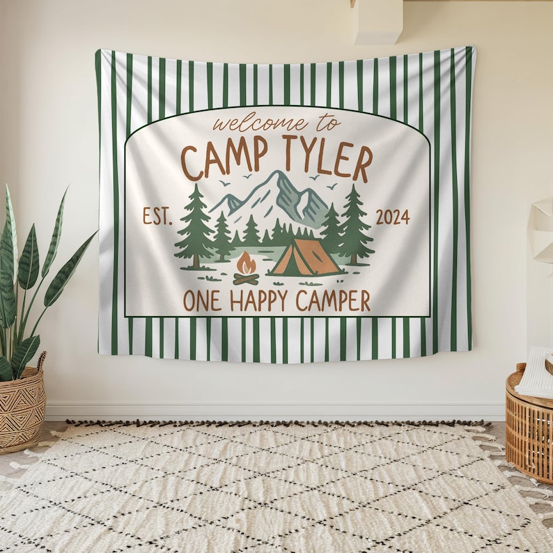 May include: A white and green striped tapestry with the text "Welcome to Camp Tyler Est. 2024 One Happy Camper". The tapestry features a mountain scene with a tent, trees, and a campfire.