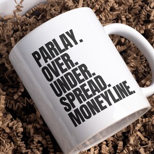 May include: White ceramic coffee mug with black text that reads "PARLAY. OVER. UNDER. SPREAD. MONEYLINE."