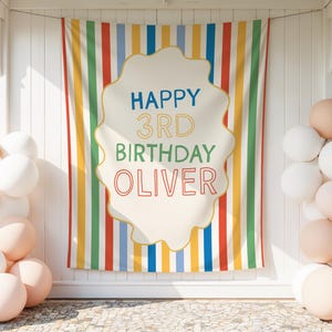 Könnte beinhalten: Ein rechteckiger Stoffhintergrund mit vertikalen Streifen in Rot, Gelb, Blau und Grün. In der Mitte befindet sich eine cremefarbene, gewellte Form mit dem Text HAPPY 3RD BIRTHDAY OLIVER in blau, gelb, grün und rot umrandeter Schrift.