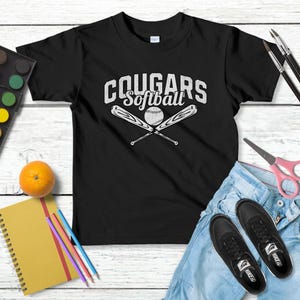 Op de afbeelding: Zwart T-shirt met de tekst "Cougars Softball" en een softbalafbeelding. De afbeelding bevat een aquarelverfset, een sinaasappel, een notitieboek, potloden, een schaar, een spijkerbroek en zwarte sneakers.