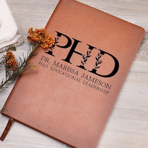 May include: A brown leather journal with the text "PhD" in black lettering with a floral design. The text "Dr. Marissa Jameson PhD, Educational Leadership" is also printed in black.