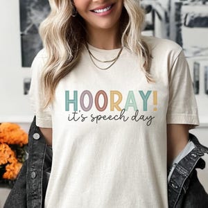Könnte beinhalten: Cremefarbenes T-Shirt mit den Worten "HOORAY! it's speech day" in bunten Buchstaben. Das Shirt hat kurze Ärmel und eine lockere Passform. Über dem Shirt wird eine dunkle Jeansjacke getragen.