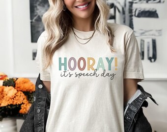 Op de afbeelding: Crèmekleurig T-shirt met de woorden "HOORAY! it's speech day" in kleurrijke letters. Het shirt heeft korte mouwen en een relaxte pasvorm. Een donker spijkerjasje wordt over het shirt gedragen.