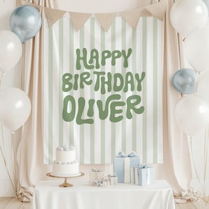 May include: A birthday party backdrop with a banner saying "Happy Birthday Oliver" in green. White and silver balloons decorate the scene. A two-tiered cake is on a gold stand, and wrapped gifts are on the table, creating a festive atmosphere.