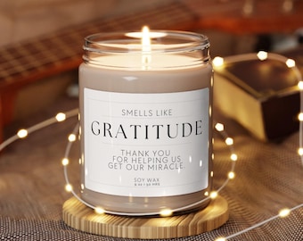 Puede incluir: Una vela encendida de cera de soja en un tarro de cristal con las palabras "Smells Like Gratitude" en una etiqueta blanca. La vela está sobre un pequeño posavasos de madera, rodeada de luces de cadena. El texto "Thank you for helping us get our miracle" también está en la etiqueta.