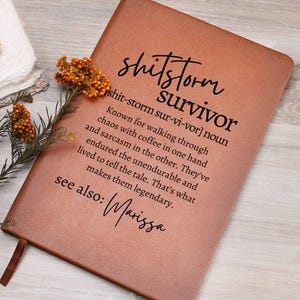 May include: A brown leather-bound journal with the words "shitstorm survivor" and a definition printed on the cover. The definition describes someone who has endured chaos with coffee and sarcasm. The name "Marissa" is also printed on the cover.