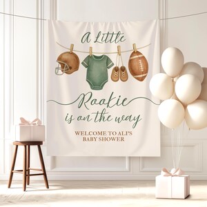 Pode incluir: Uma faixa de ch&aacute; de beb&eacute; com o texto "a little Rookie is on the way". A faixa apresenta ilustra&ccedil;&otilde;es de um capacete de futebol americano, um body, sapatos de beb&eacute; e uma bola de futebol americano. Bal&otilde;es brancos e uma caixa de presente tamb&eacute;m est&atilde;o presentes.