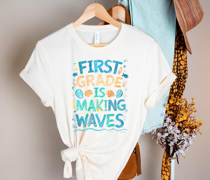 Pode incluir: Uma camiseta branca com um gr&aacute;fico azul e amarelo que diz "First Grade is Making Waves". O gr&aacute;fico inclui conchas e peixes.