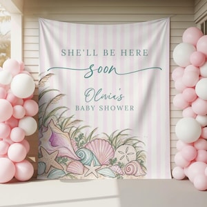 May include: A fabric backdrop featuring vertical light pink and white stripes with the text SHE'LL BE HERE soon Olivia's BABY SHOWER. The bottom left corner displays a watercolor illustration of various seashells, starfish, and beach grass.