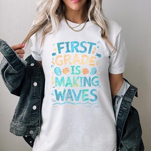 Pode incluir: Camiseta branca com um gr&aacute;fico colorido que diz "First Grade is Making Waves". O gr&aacute;fico inclui conchas, peixes e ondas.