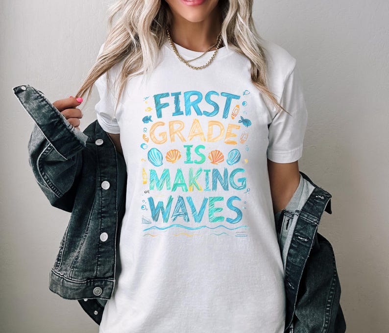 Pode incluir: Camiseta branca com um gr&aacute;fico colorido que diz "First Grade is Making Waves". O gr&aacute;fico inclui conchas, peixes e ondas.