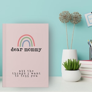 May include: A pink journal with the text "dear mommy" and a rainbow illustration. Below the title, the text reads "all the things I want to tell you". The journal is on a white surface with other decorative items.
