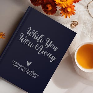 May include: A navy blue journal with the text "While You Were Away" and "memories to share when we reunite" in silver lettering. A heart symbol is also included in the silver lettering.