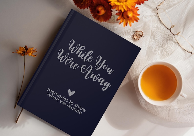 May include: A navy blue journal with the text "While You Were Away" and "memories to share when we reunite" in silver lettering. A heart symbol is also included in the silver lettering.