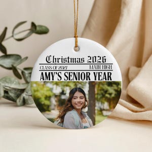 May include: A round white ceramic ornament hanging from a gold string. The text reads Christmas 2026, Class of 2021, Main High, and Amy's Senior Year. Below the text is a photographic portrait of a person with long brown hair smiling outdoors.