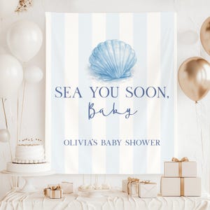 May include: A baby shower banner with blue and white stripes, a watercolour seashell, and the text "SEA YOU SOON, Baby, Olivia's Baby Shower." White and gold balloons, a cake, and gifts are also visible.