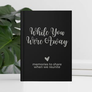 May include: Black journal with the text "While You Were Away" in silver lettering. A small heart symbol is below the text, and the words "memories to share when we reunite" are below the heart.