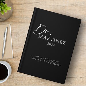 Puede incluir: Diario de tapa dura negro con texto blanco que dice "Dr. Martinez 2024" y "Ph.D. Education University of Miami".