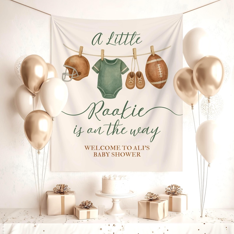 Pode incluir: Cena de ch&aacute; de beb&eacute; com uma faixa que diz "A Little Rookie is on the way". A faixa apresenta ilustra&ccedil;&otilde;es em aquarela de um capacete de futebol americano, um body, sapatos de beb&eacute; e uma bola de futebol. Bal&otilde;es dourados e brancos, presentes e um bolo.
