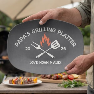 Puede incluir: Una fuente ovalada para asar a la parrilla de color gris oscuro con el texto "PAPA'S GRILLING PLATTER" en blanco. La fuente presenta un tenedor y una espátula cruzados, un gráfico de llama y el texto "LOVE, NOAH & ALEX".