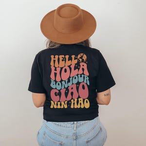 May include: Black t-shirt with the words "HELLO HOLA BONJOUR CIAO NIN HAO" in a retro font. The shirt has a globe graphic and is worn with a brown hat and blue jeans.