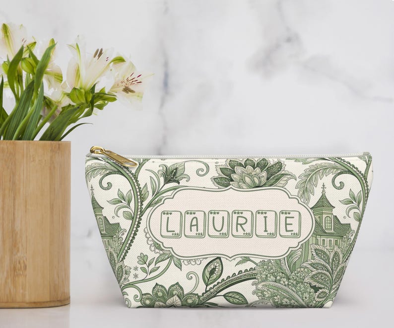 K&ouml;nnte beinhalten: Eine wei&szlig;e Kosmetiktasche mit gr&uuml;nem Blumen- und Architekturmuster. Die Tasche hat einen goldenen Rei&szlig;verschluss und den Namen "LAURIE" auf der Vorderseite. Links steht eine Bambusvase mit wei&szlig;en Blumen.